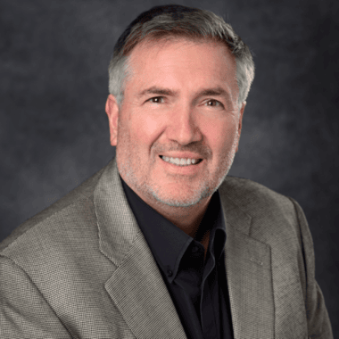 Christopher Contag smiles at the camera. He is sitting for a formal portrait and is wearing a gray suit with black shirt. He has close-cut slightly graying hair and a very slight beard and mustache. 